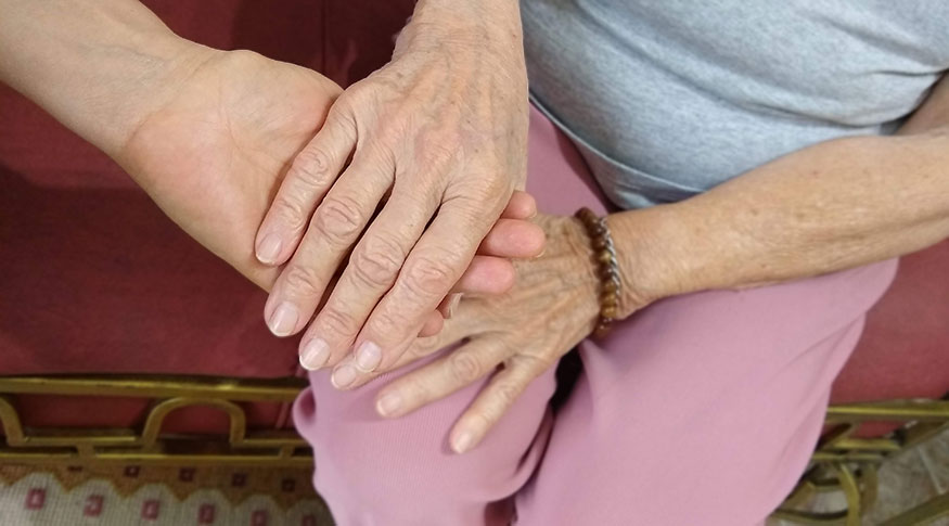 #PraCegoVer A foto mostra uma mão de uma pessoa adulta entrelaçada na mão de uma pessoa idosa, apoiadas no colo da pessoa idosa